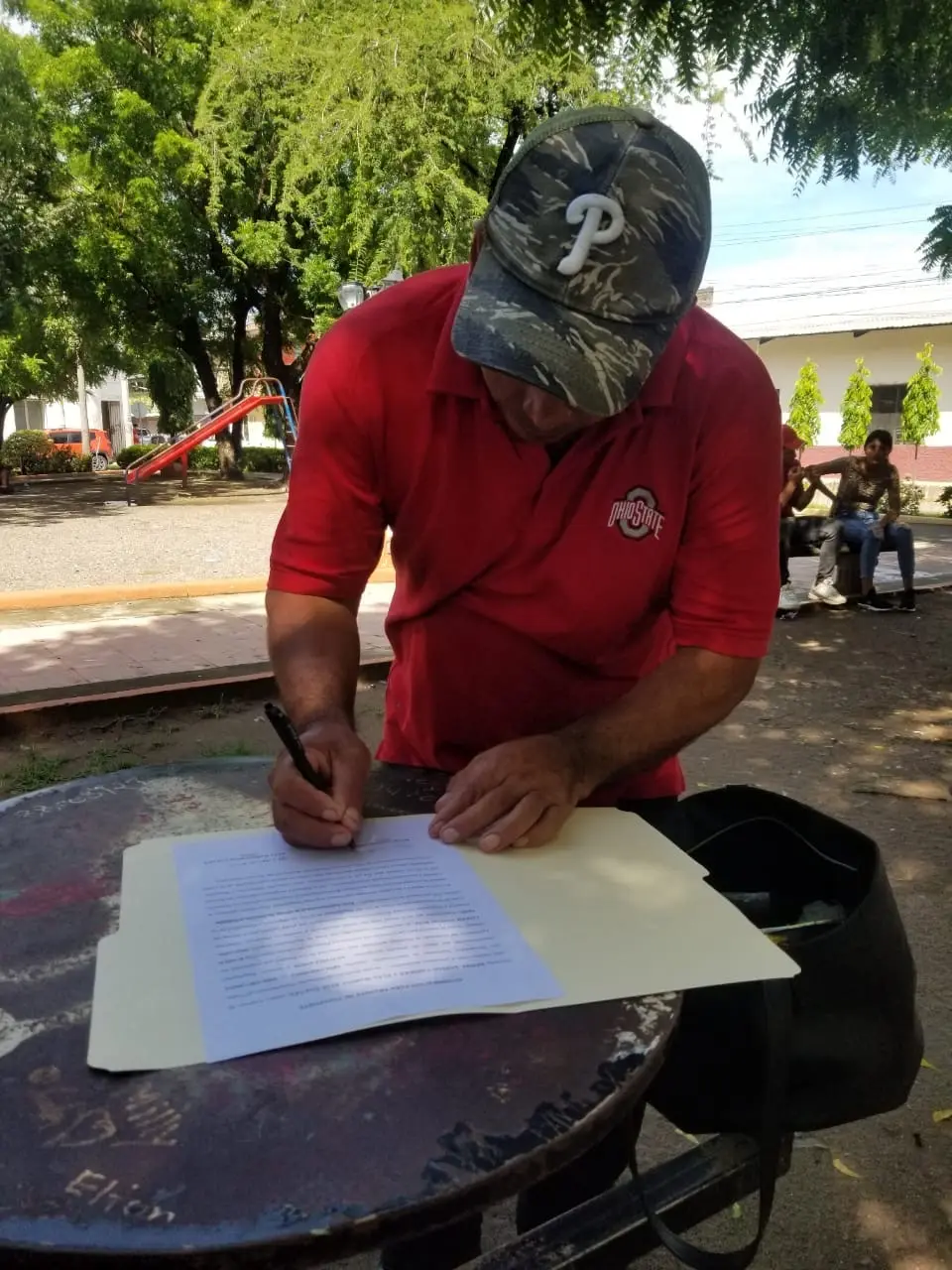 Cliente hondureño firma documentos legales sobre una mesa en un parque de Honduras con acompañamiento de Abogados Catrachos USA.